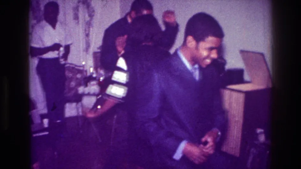 1976 Harlem indoor party frame with a suited man smiling in the foreground, dancers behind him, and a bottle near the back wall.