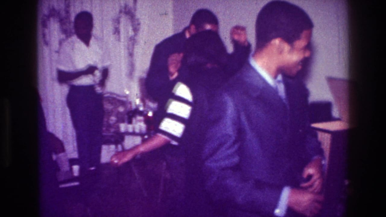1976 Harlem home-movie frame showing Black men at an indoor party, with a smiling suited man in front and guests near a drink table.