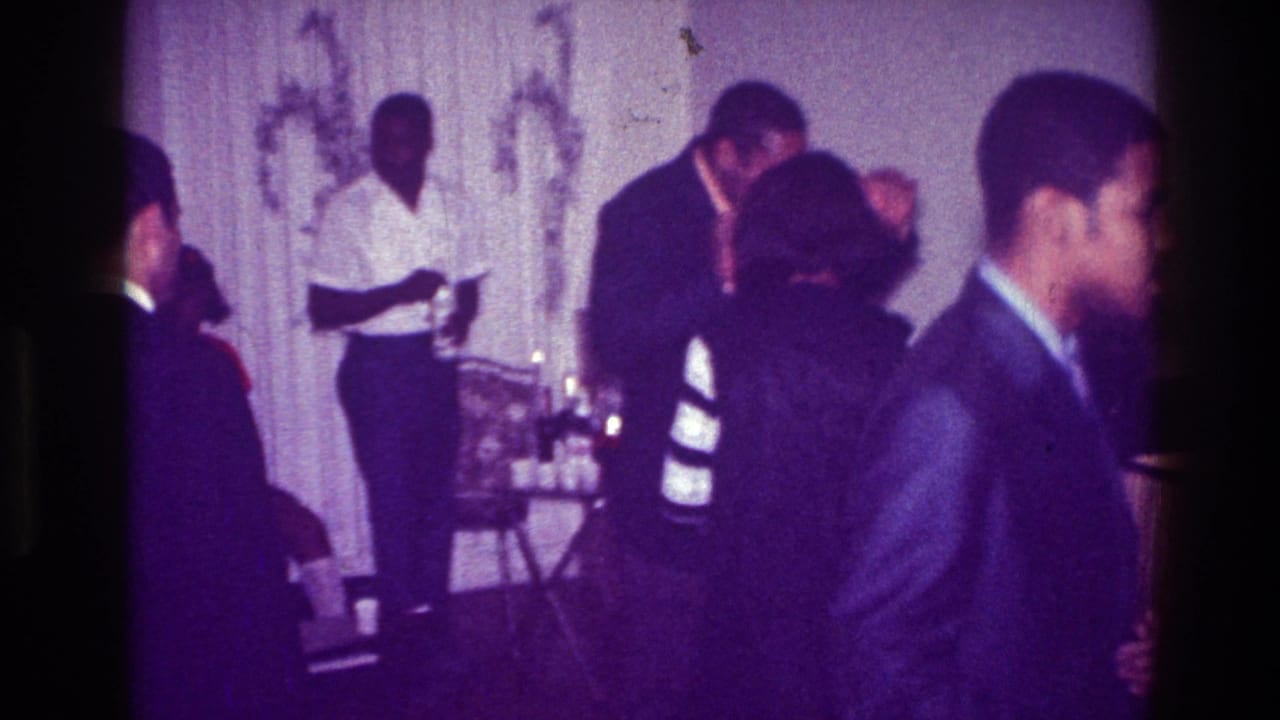 1976 Harlem party frame with men in suits and formal wear moving through a private interior near a small drink table.
