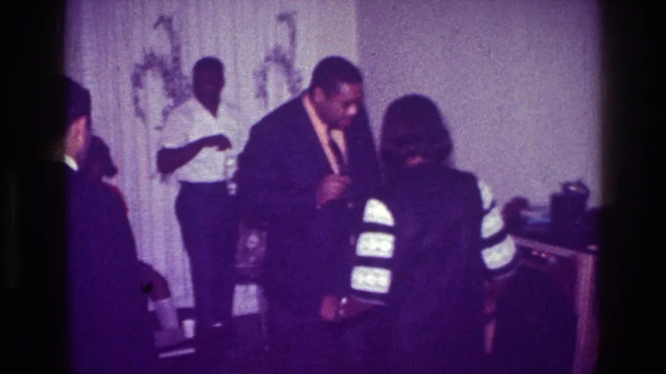 1976 Harlem indoor party frame showing suited guests interacting closely in a dim private room with curtains and chairs.