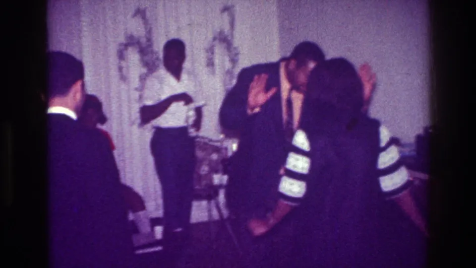 1976 Harlem home-movie frame with a suited man raising his hand during a lively indoor party in a private interior.