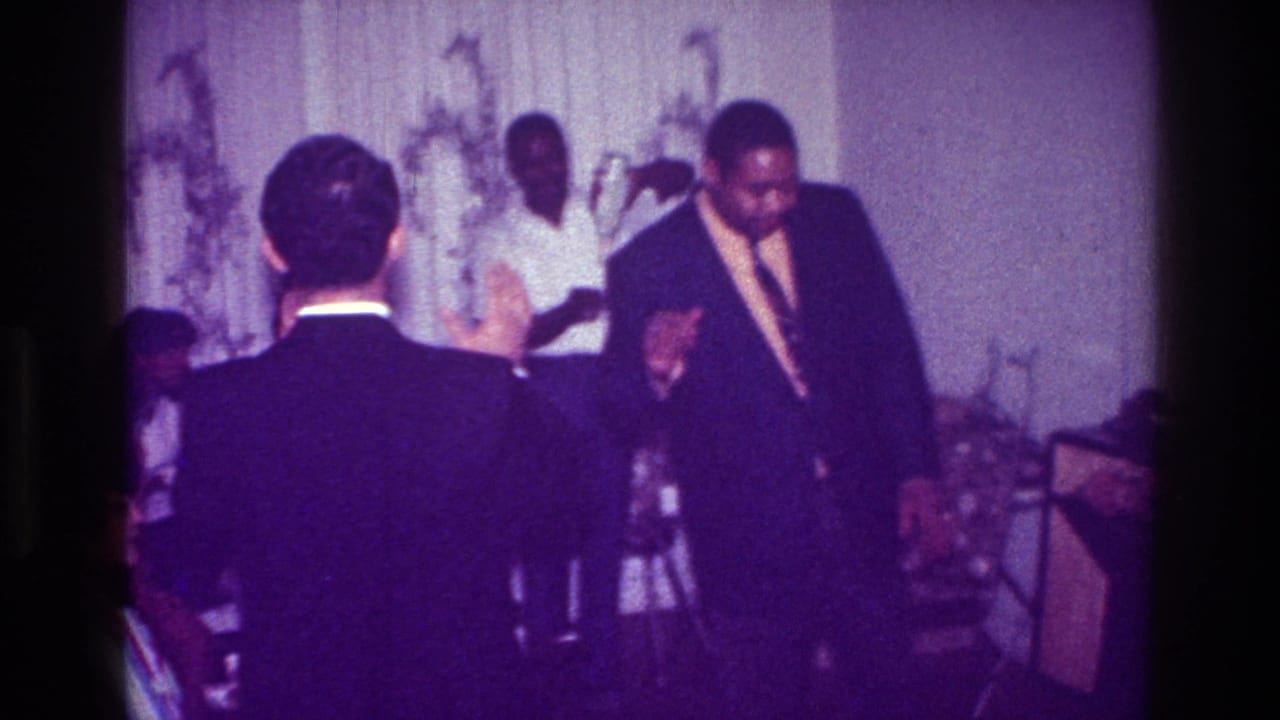 In a 1976 Harlem home-movie frame, a man in a dark suit dances in front of patterned curtains as other guests watch nearby.
