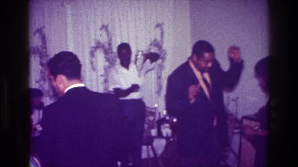 A foreground guest looks down or turns aside while the suited dancer continues moving in the private Harlem gathering.