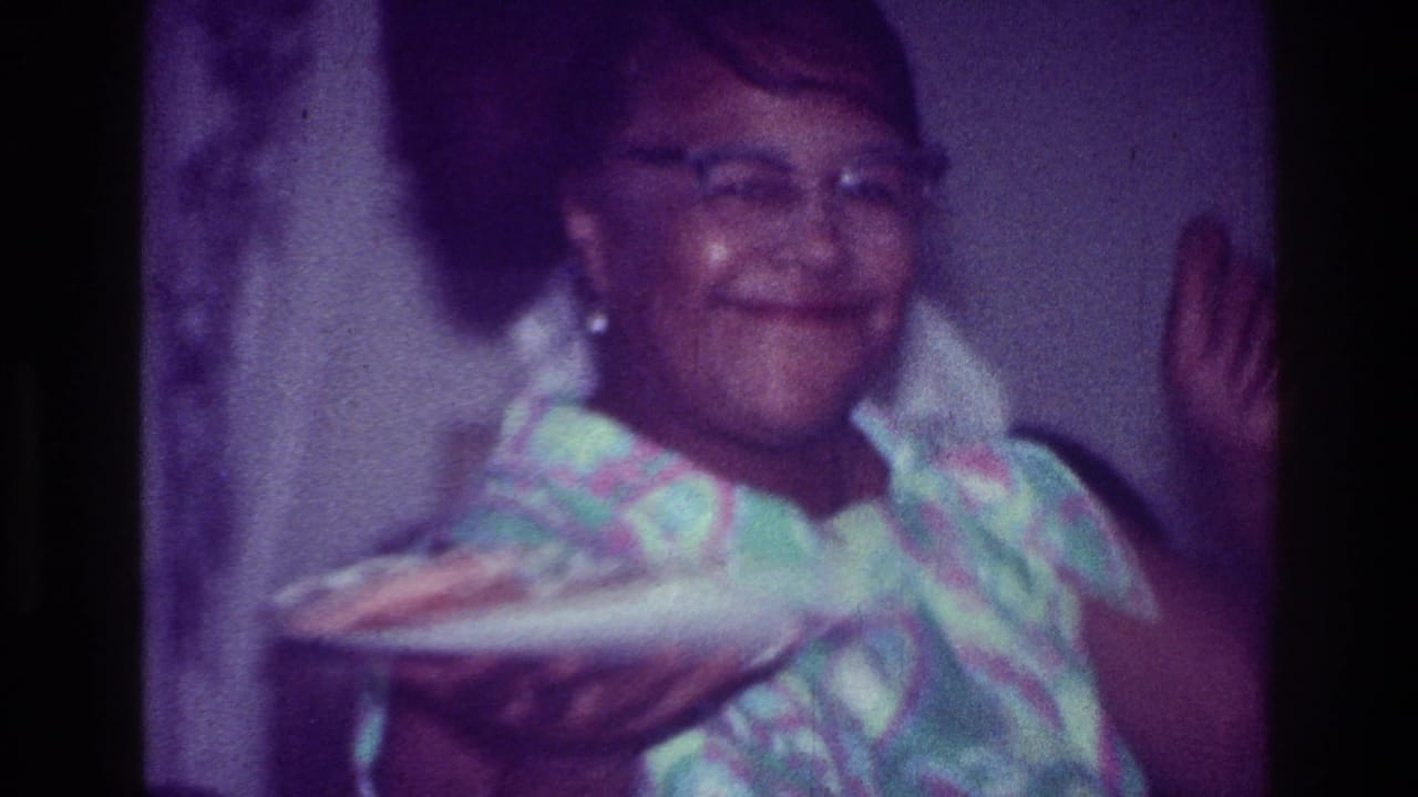 In supported 1976 Harlem home-movie footage, a smiling woman in glasses holds a plate while standing in a private interior.