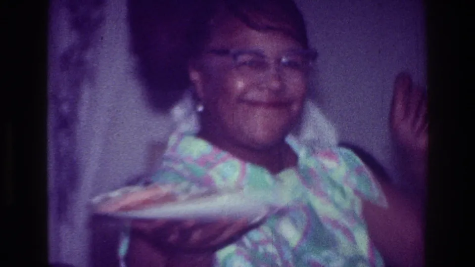 In supported 1976 Harlem home-movie footage, a smiling woman in glasses holds a plate while standing in a private interior.