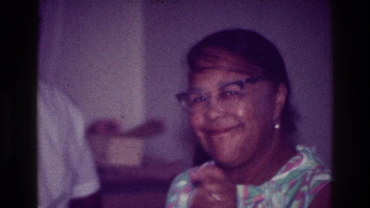 Woman in glasses smiling and gesturing with a cup or hand near a table.