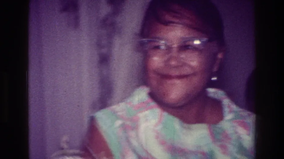 Close 8mm frame of the woman smiling toward the camera in glasses and a patterned dress inside the private residence.