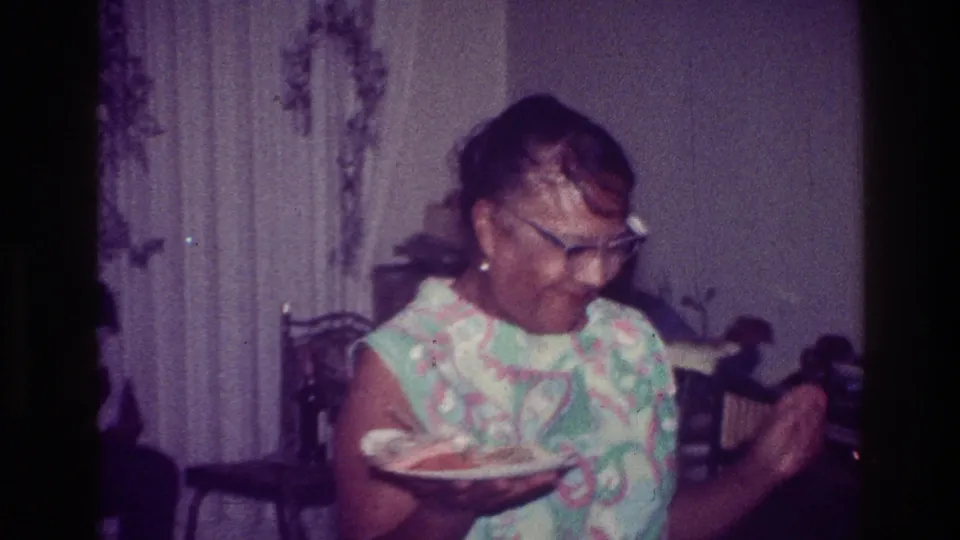 The woman dances while holding a plate in a private Harlem interior, with curtains and chairs visible behind her.