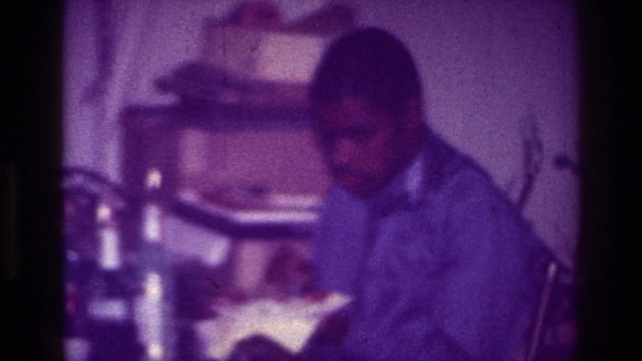A Black man in a collared shirt sits at a table in a Harlem interior, eating from a bowl amid household clutter.