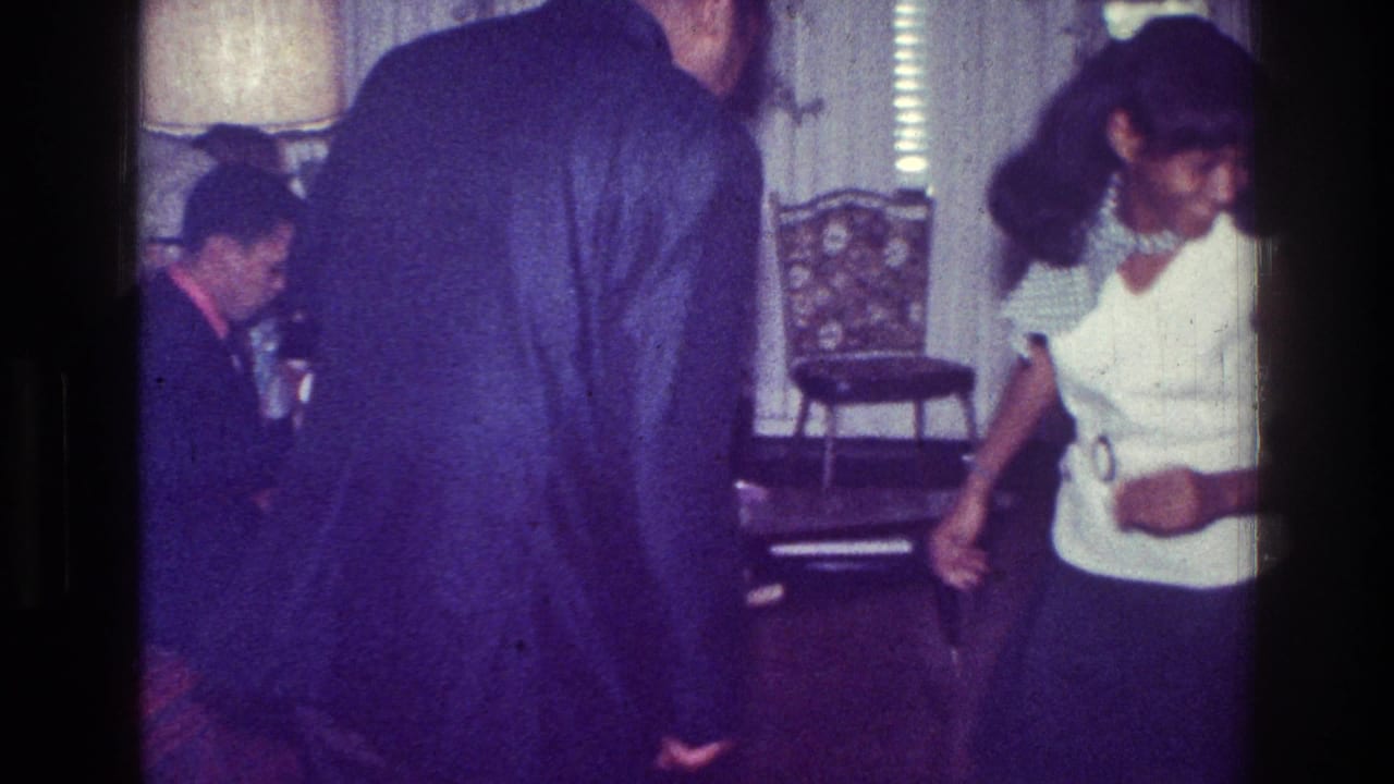 1976 Harlem home-movie frame showing adults in a living room, with a woman in a white top moving near furniture and patterned wallpaper.