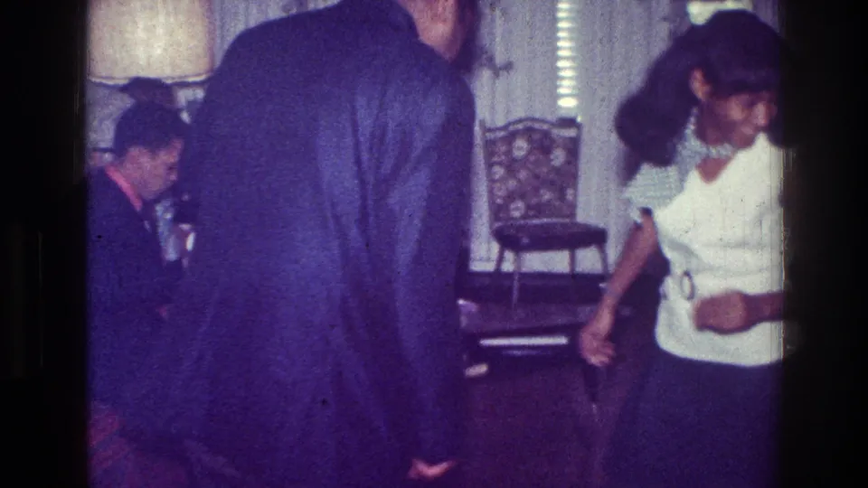 1976 Harlem home-movie frame showing adults in a living room, with a woman in a white top moving near furniture and patterned wallpaper.