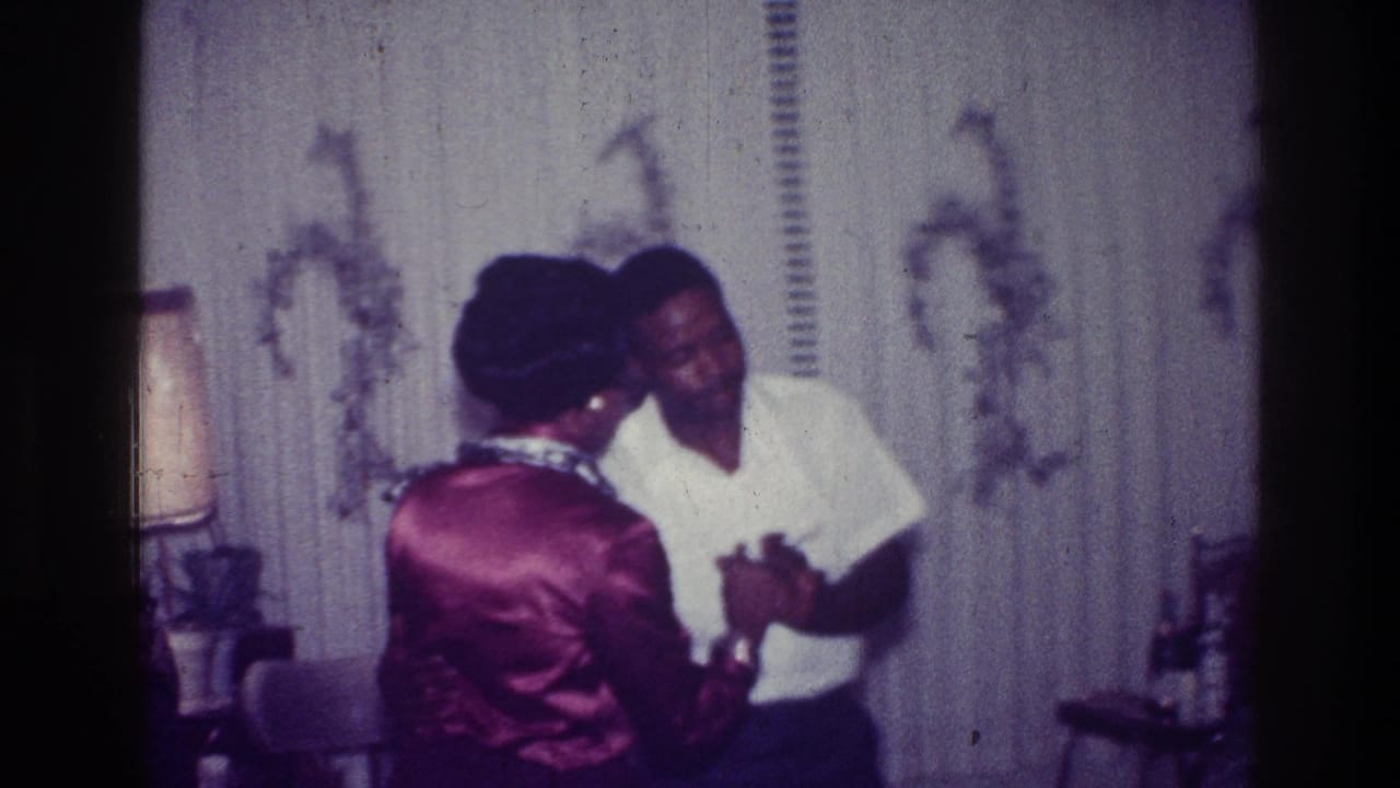 1976 Harlem frame of a couple dancing closely indoors, with a table lamp, patterned wallpaper, and home furnishings visible.