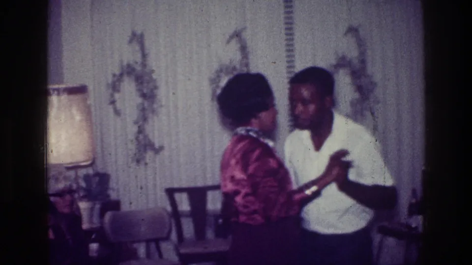 1976 Harlem home-movie frame showing a couple dancing in a private living room with chairs, a lamp, and patterned wallpaper.