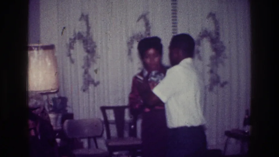 1976 Harlem frame of a couple dancing in profile inside a living room, with period wallpaper, chairs, and lamp visible.