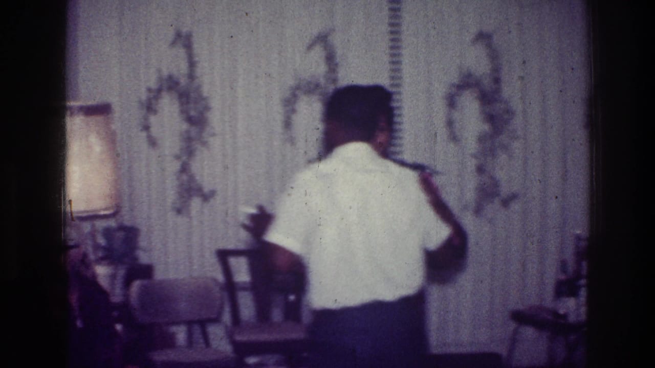 1976 Harlem home-movie frame showing a man in a white short-sleeved shirt dancing inside a decorated living room.