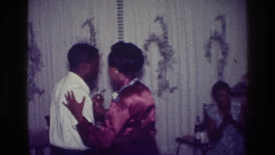 1976 Harlem home-movie frame showing a couple dancing closely while a seated guest watches from the background.