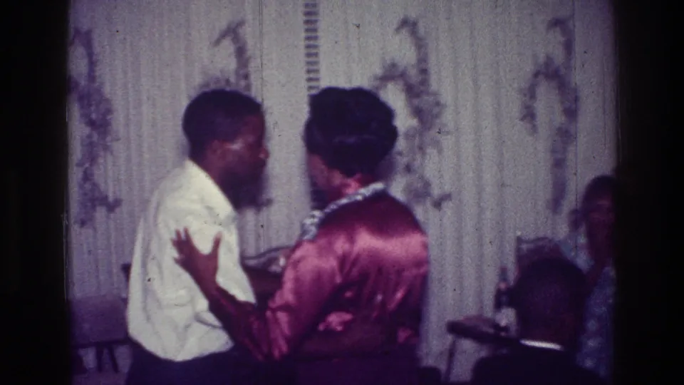1976 Harlem home-movie frame of a couple dancing face to face in a living room with guests seated nearby.