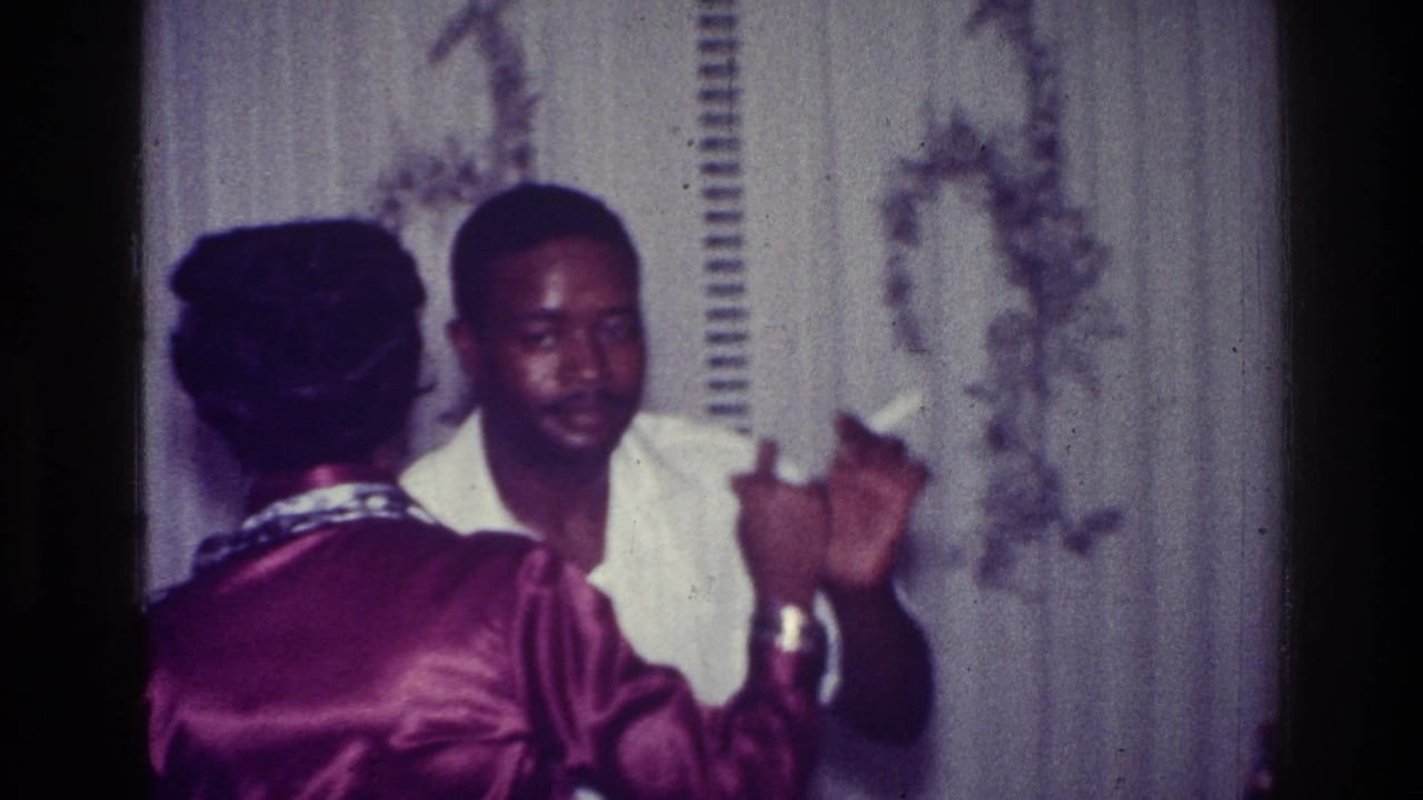 A Black man in a white collared shirt dances with a woman in a magenta satin-like dress against light patterned curtains in 1976 Harlem footage.