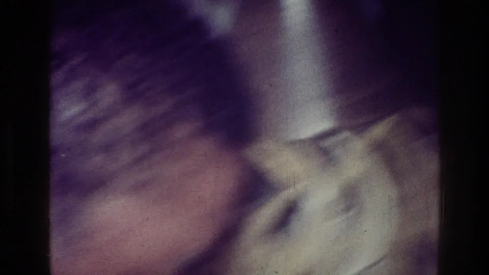 1976 Harlem home-movie frame showing a blurred close view of a person in a light shirt within a shadowed private interior.