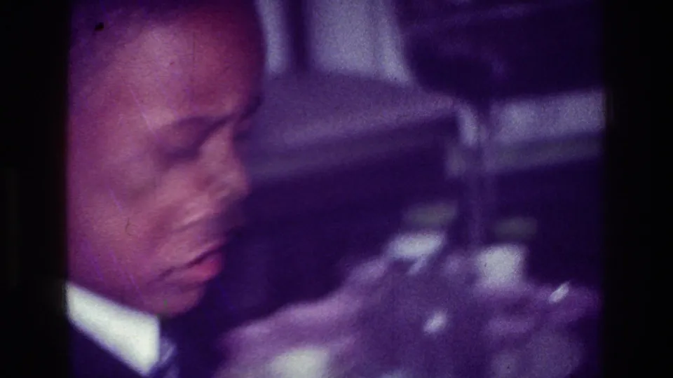 1976 Harlem home-movie frame with a close-up of an adult man seated in a dim private interior, his face turned downward.