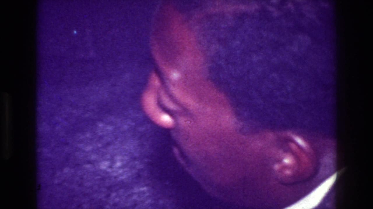 Close handheld 1976 Harlem home-movie frame showing a person’s head and shoulder in a dim party interior with heavy purple color shift.