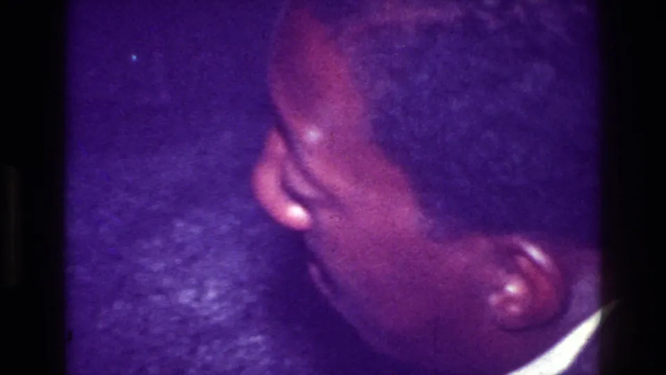 Close handheld 1976 Harlem home-movie frame showing a person’s head and shoulder in a dim party interior with heavy purple color shift.