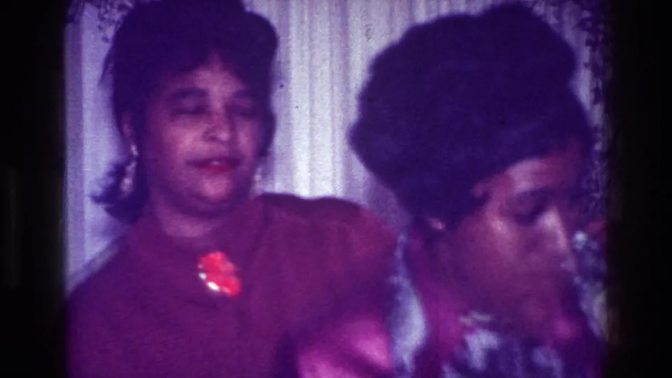 Two young women at a 1976 Harlem home party stand near patterned curtains, with one in a purple top and red brooch as they appear to dance or react.