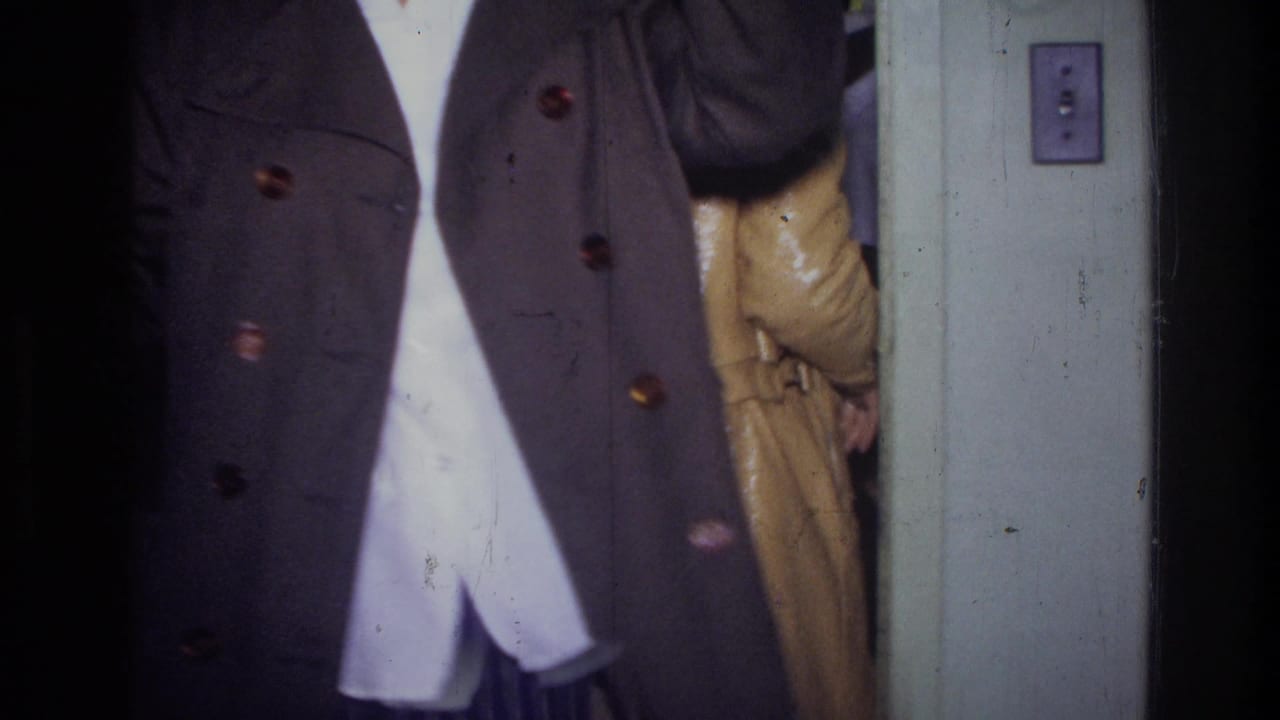 1976 Harlem home-movie frame showing a cropped figure in a dark coat beside a doorway, with another coat partly visible behind.
