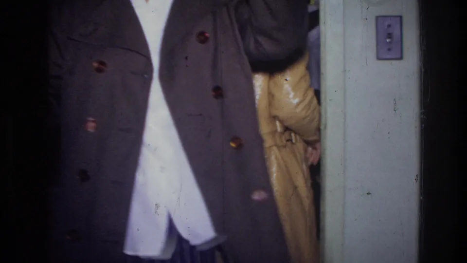 1976 Harlem home-movie frame showing a cropped figure in a dark coat beside a doorway, with another coat partly visible behind.