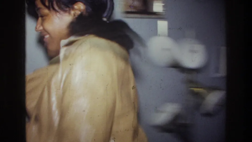1976 Harlem home-movie frame showing a smiling young woman in a tan coat in profile inside a private interior.