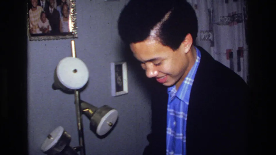 1976 Harlem home-movie frame showing a young man in a dark jacket and blue patterned shirt near a wall lamp and framed photo.