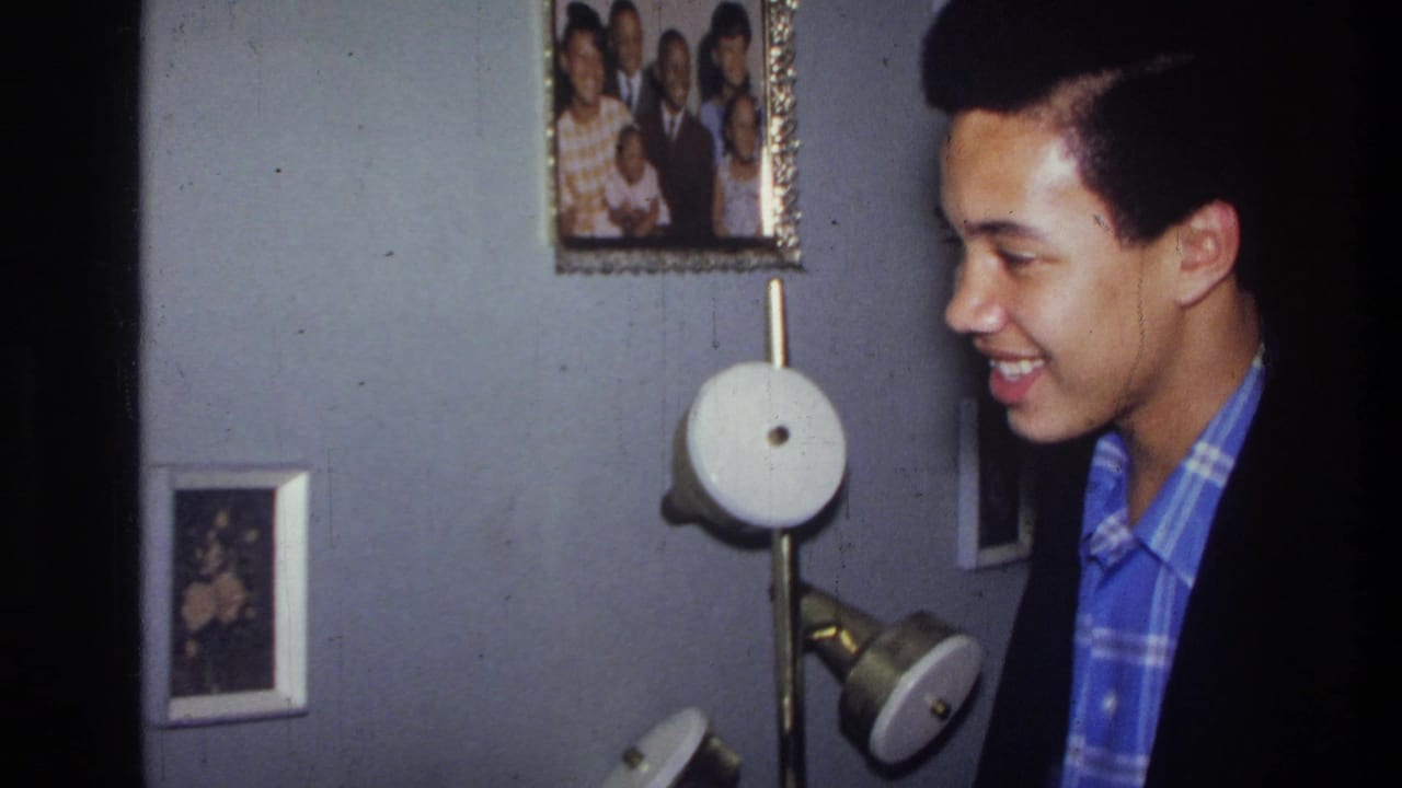 Young man in a blue plaid shirt smiles in an apartment hallway near framed photos and a wall lamp.
