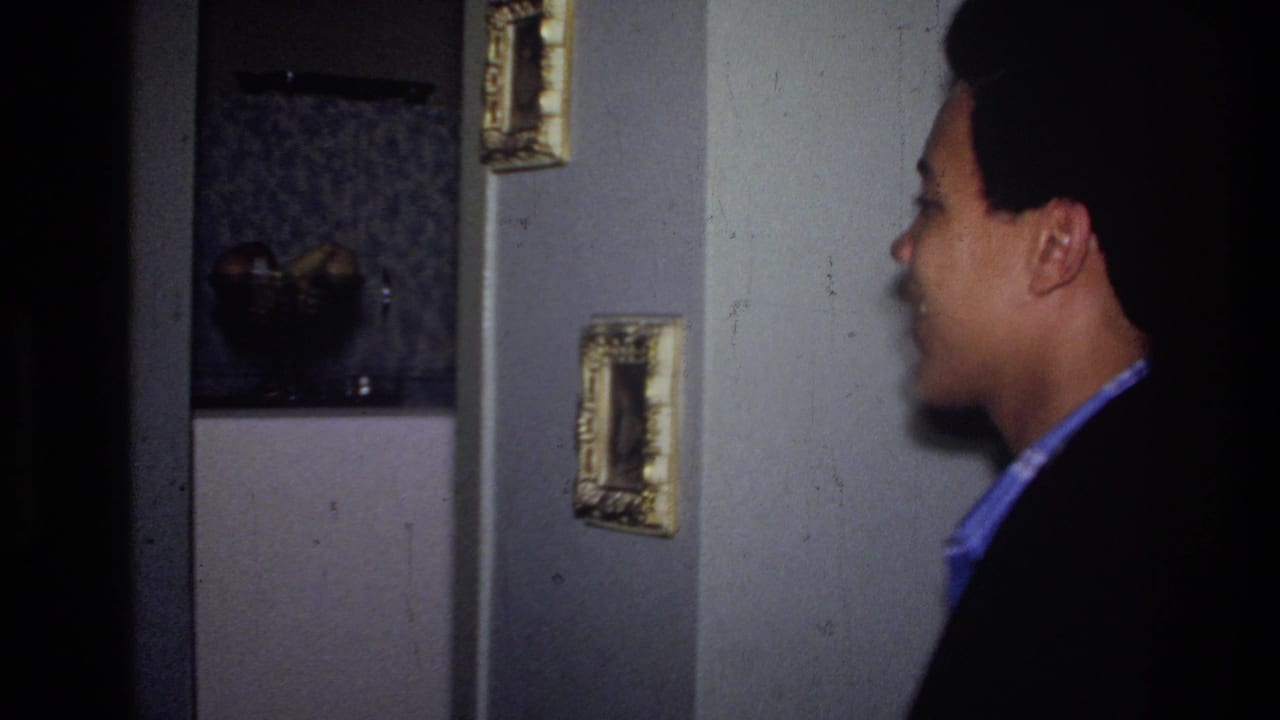 The young man appears in profile inside the Harlem apartment, with framed pictures, a doorway, and a darker room behind him.