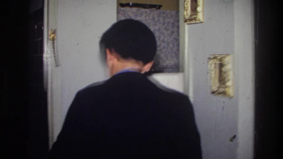 From behind, the young man moves through a Harlem apartment doorway, with patterned wallpaper and framed wall decor nearby.