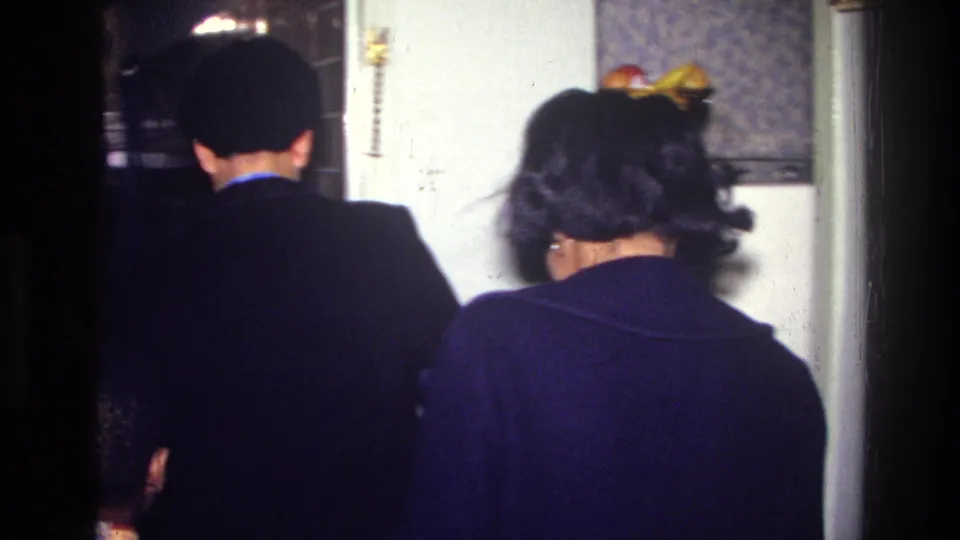 Two people move away from the camera inside the 1976 Harlem apartment, near patterned wallpaper, a doorway, and a fruit bowl.