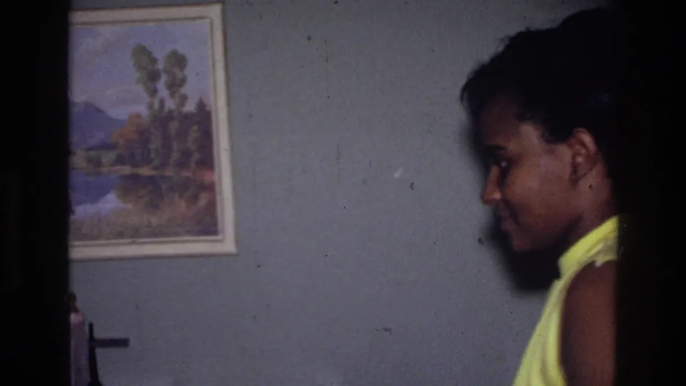 In a 1976 Harlem home-movie frame, a woman in a yellow sleeveless dress stands indoors in profile near framed wall art.