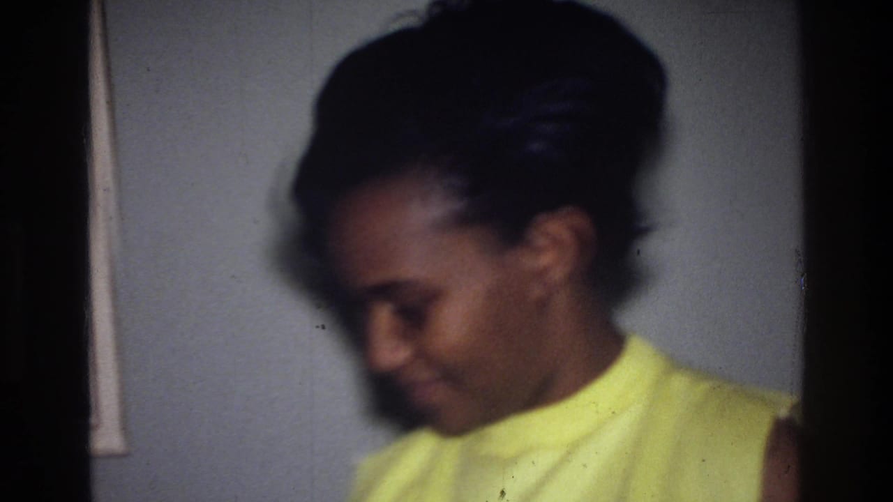 The woman appears softly focused as she looks down indoors, with the yellow dress standing out against the muted wall.
