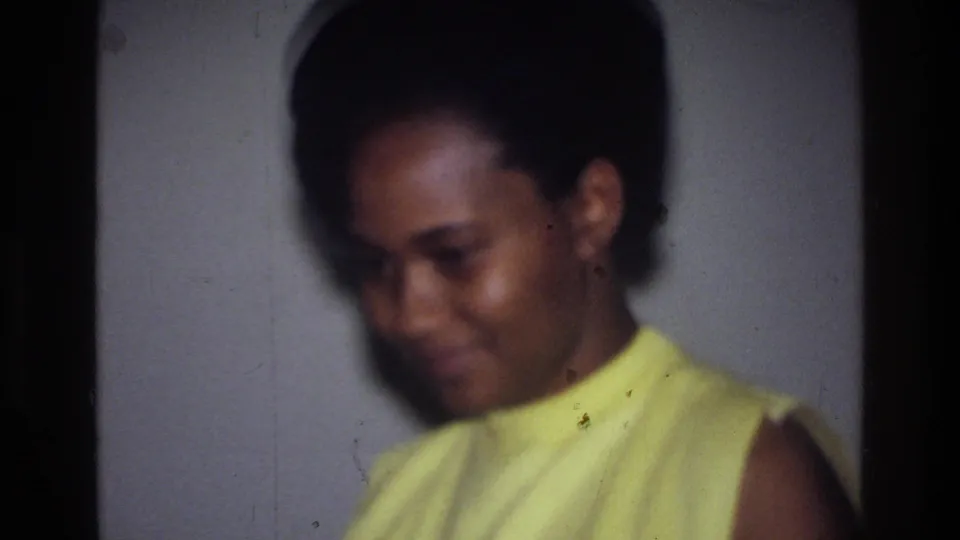 In the Harlem home-movie setting, the woman smiles faintly while looking down, with soft focus and grain visible.