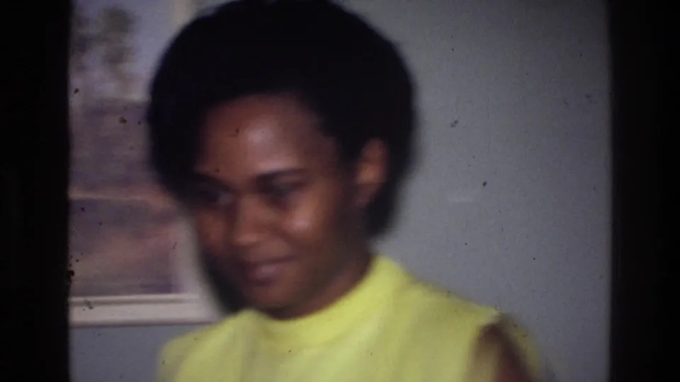 The woman faces closer toward the camera in this 1976 Harlem interior frame, with framed wall art partly visible behind her.