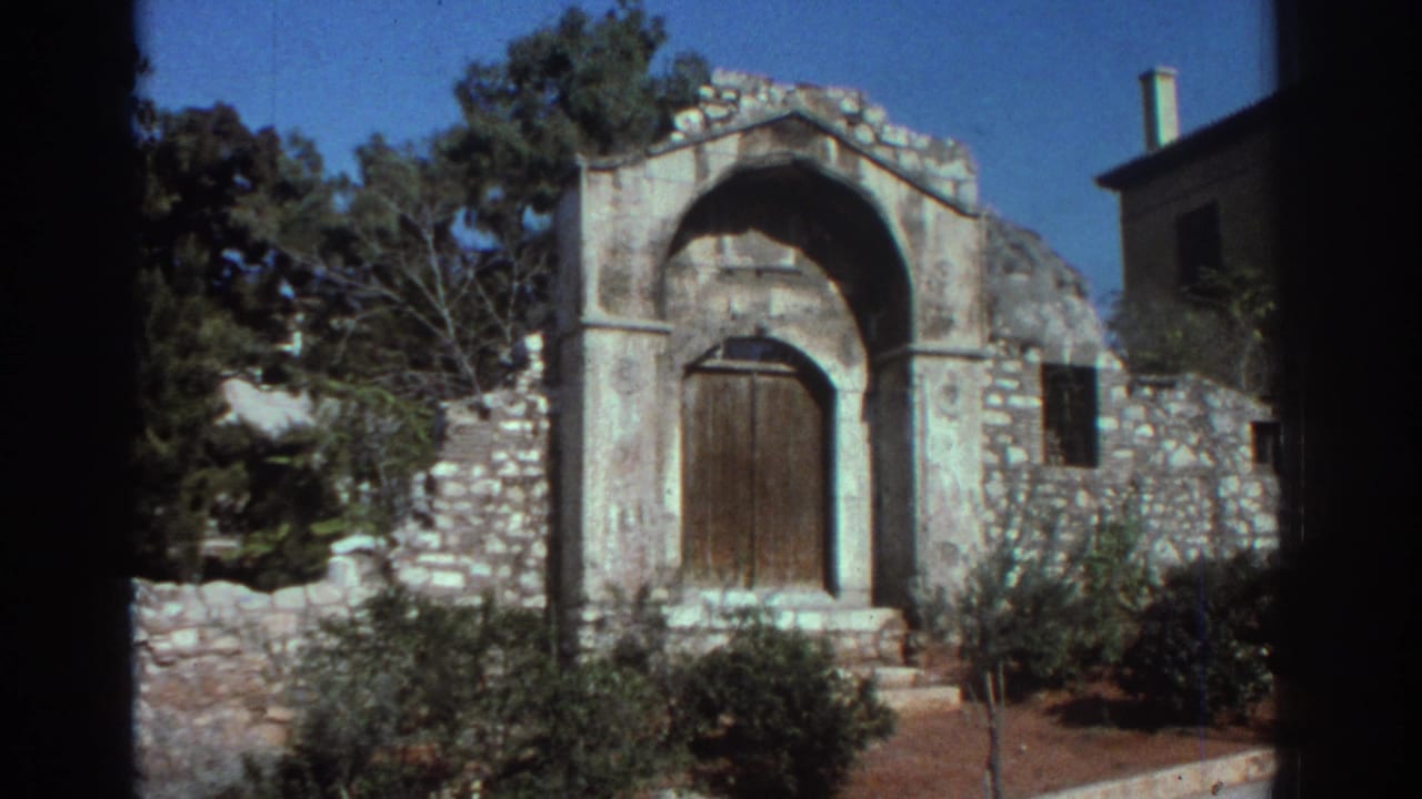 Stone church entrance with arched wooden door, low wall, trees, and shrubs under a blue sky.