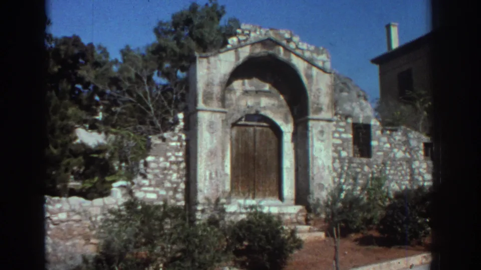 1976 Tel Aviv 8mm frame showing a weathered stone church entrance with an arched wooden door, surrounding wall, trees, and shrubs.