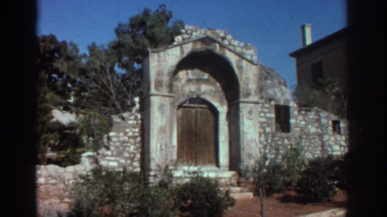 Weathered arched doorway in a stone structure with greenery and a nearby building edge.