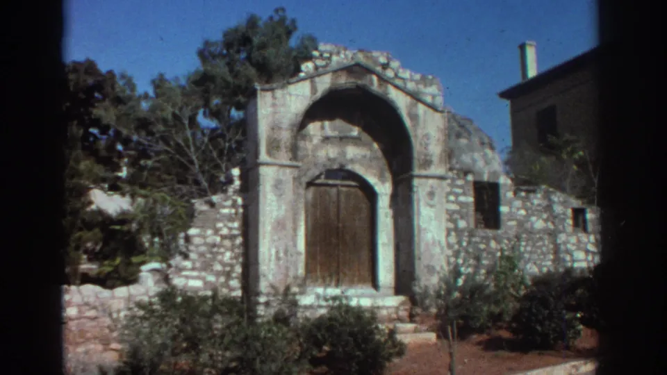 1976 Tel Aviv travel frame of a stone religious structure with an arched wooden door, rough masonry, shrubs, and a building edge nearby.
