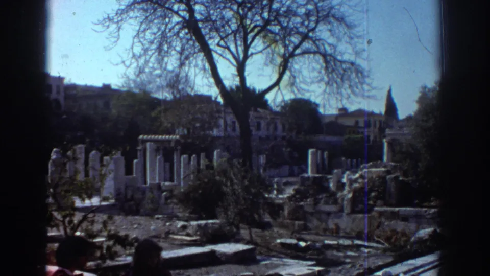 1976 Tel Aviv 8mm frame looking across archaeological ruins, columns, stone fragments, a leafless tree, distant buildings, and partial visitors.