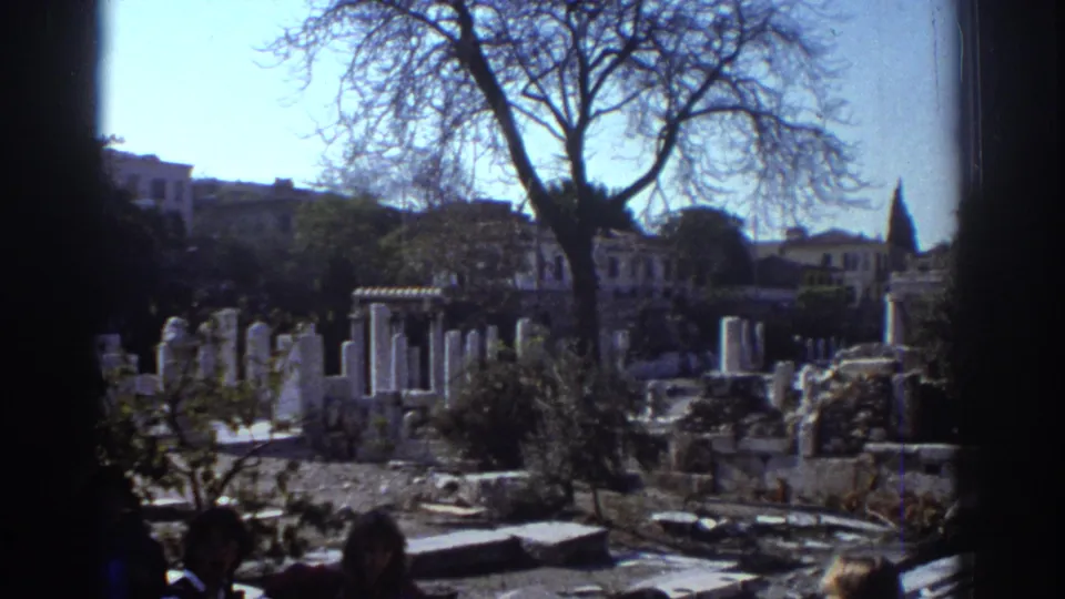 1976 Tel Aviv home-movie frame of ruins and white columns seen past a leafless tree, with distant buildings and indistinct visitors nearby.