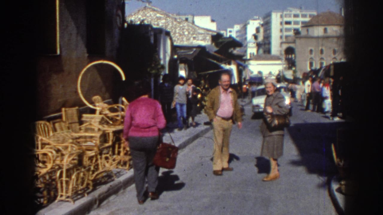 Pedestrians walk along a narrow market street beside stacked wicker furniture.