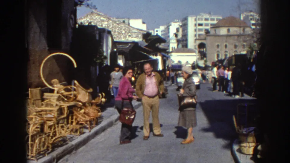 1976 Tel Aviv frame showing three adults near stacked wicker furniture, with market awnings, cars, and mixed buildings behind them.