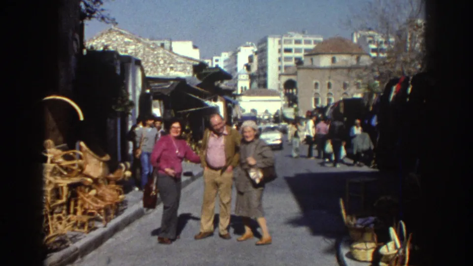 1976 Tel Aviv street-market frame with three adults walking toward the camera beside wicker furniture, awnings, cars, and background buildings.