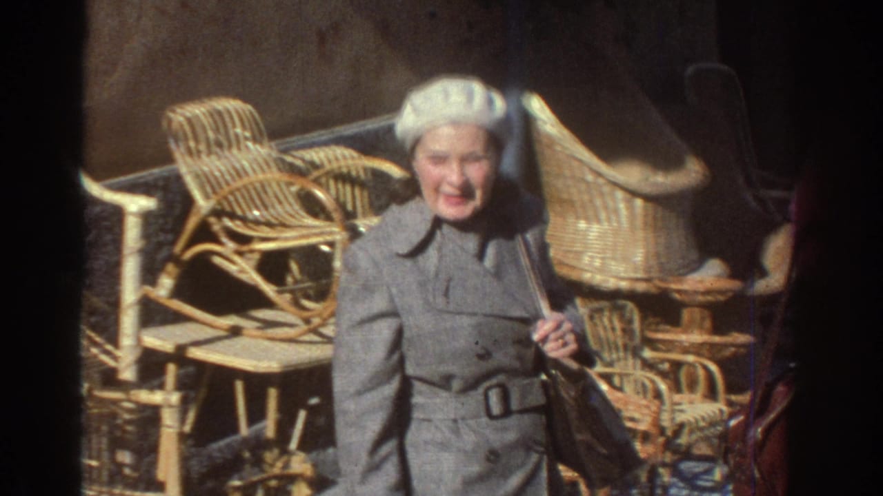1976 Tel Aviv home-movie frame showing a woman in a grey coat and white beret beside wicker chairs in outdoor light.