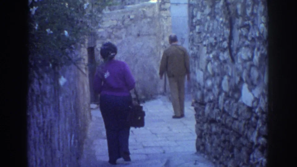 1976 Tel Aviv home-movie frame with two adults seen from behind in a cool-toned stone alley.